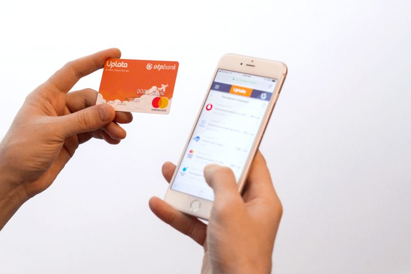 Hand holding an Uplata and OTP Bank Mastercard, next to a phone with the Uplata app
